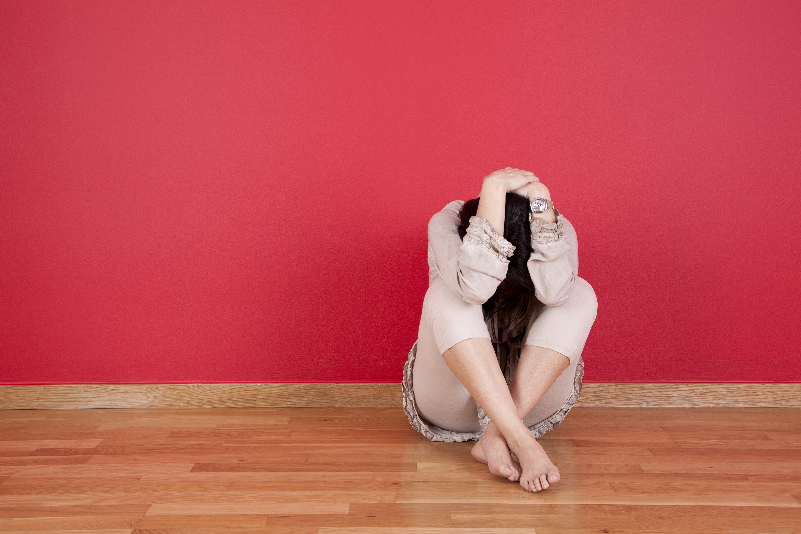 An image of a woman sitting and reclining against a red wall, who is holding her head in her hands